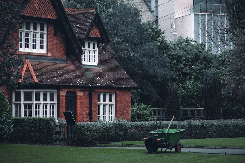 A red brick house with a steep, tiled roof and white-framed windows is situated in a garden. The garden features neatly trimmed hedges, a well-maintained grassy lawn, and a variety of trees and shrubs