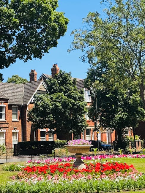 A red brick house with a steep, tiled roof and white-framed windows is situated in a garden. The garden features neatly trimmed hedges, a well-maintained grassy lawn, and a variety of trees and shrubs